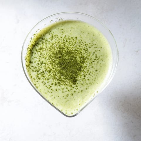 A creamy Matcha Martini cocktail beside a matcha tin, showcasing a smooth, pale green beverage in a stemmed glass.  